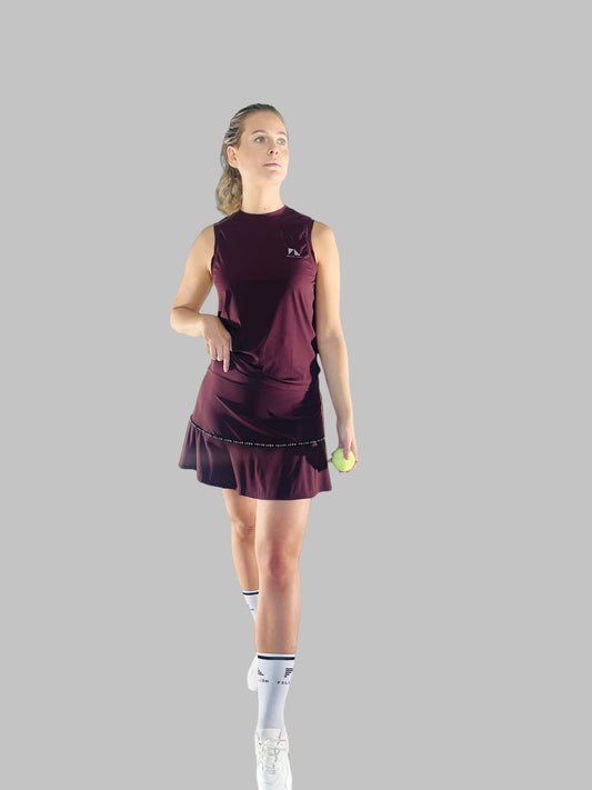 Full kroppsbild av modell klädd i ett matchande sportset med Iza Performance Skirt och en topp i burgundy, poserar på en enfärgad bakgrund, perfekt utrustad för både padel och tennis.