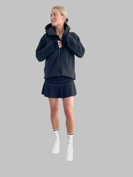 Modell som bär en outfit, uppvärmningströja, padelkjol och sportstrumpor. 